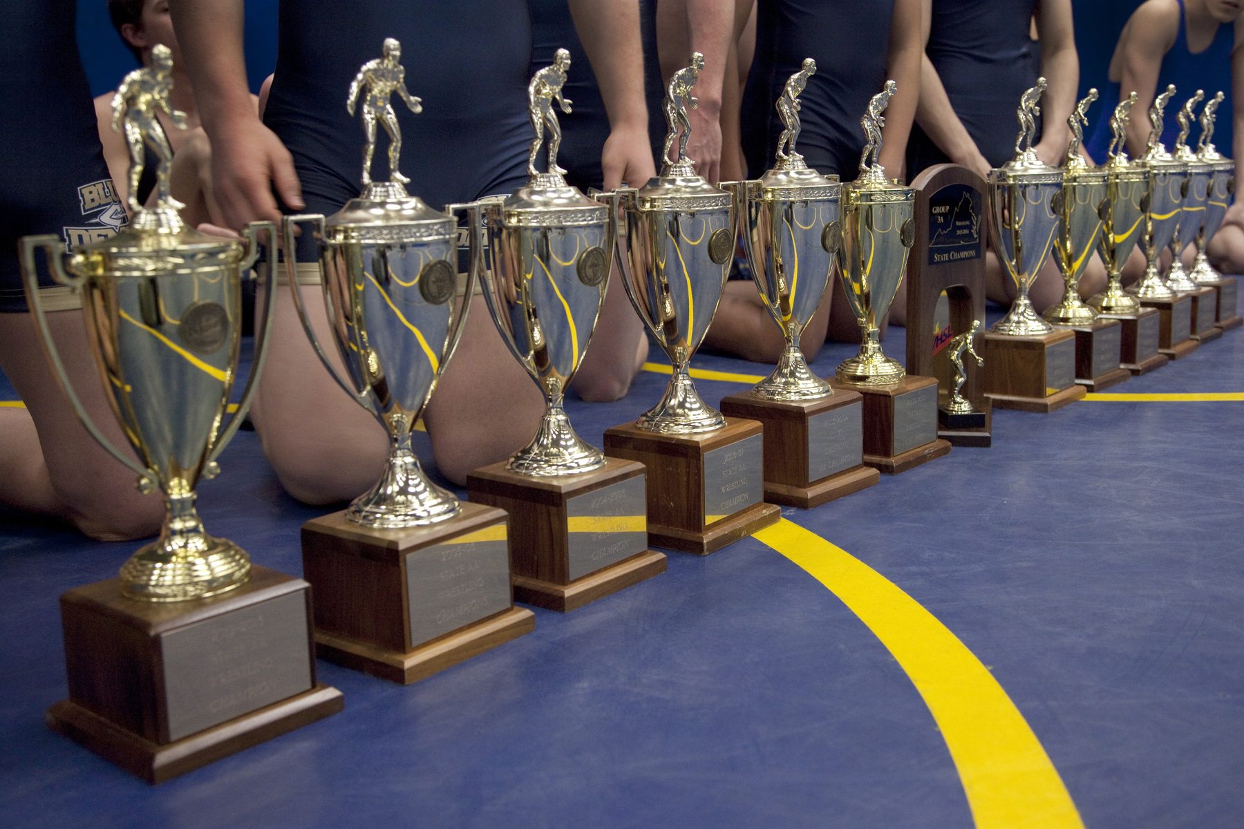Row of team state championship trophies lined up on the wrestling mat, team kneeling behind them